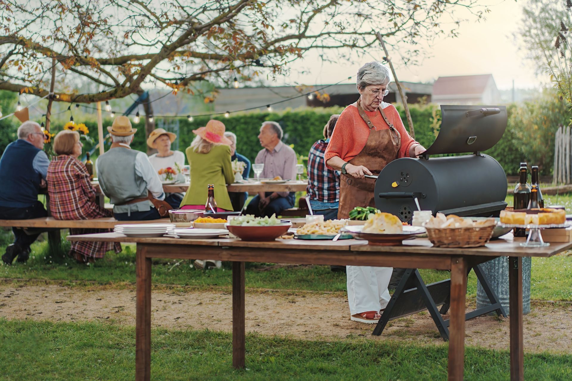 Senior Woman Barbecuing at Countryside Gathering - A senior woman prepares food at a barbecue as her friends chat under a lit tree at sunset.