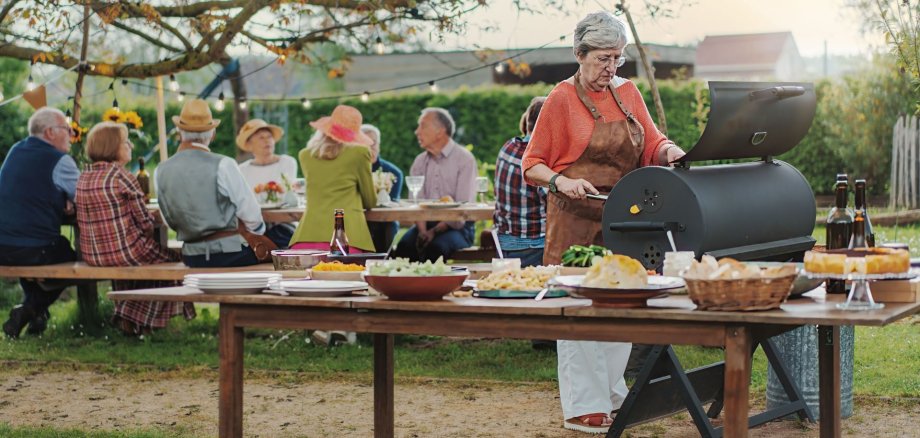 Senior Woman Barbecuing at Countryside Gathering - A senior woman prepares food at a barbecue as her friends chat under a lit tree at sunset.